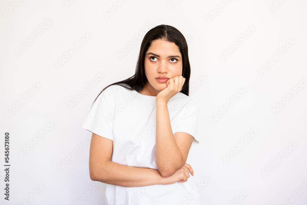 Portrait of thoughtful or bored young woman posing with hand on chin over white background. Caucasian lady wearing white T-shirt standing with pensive expression. Thinking or planning concept Portrait of thoughtful or bored young woman posing with hand on chin over white background. Caucasian lady wearing white T-shirt standing with pensive expression. Thinking or planning concept
