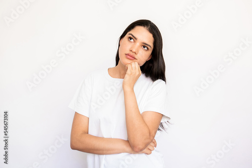 Portrait of confident young woman thinking over idea. Caucasian lady wearing white T-shirt standing with hand on chin and looking away over white background. Thinking or planning concept