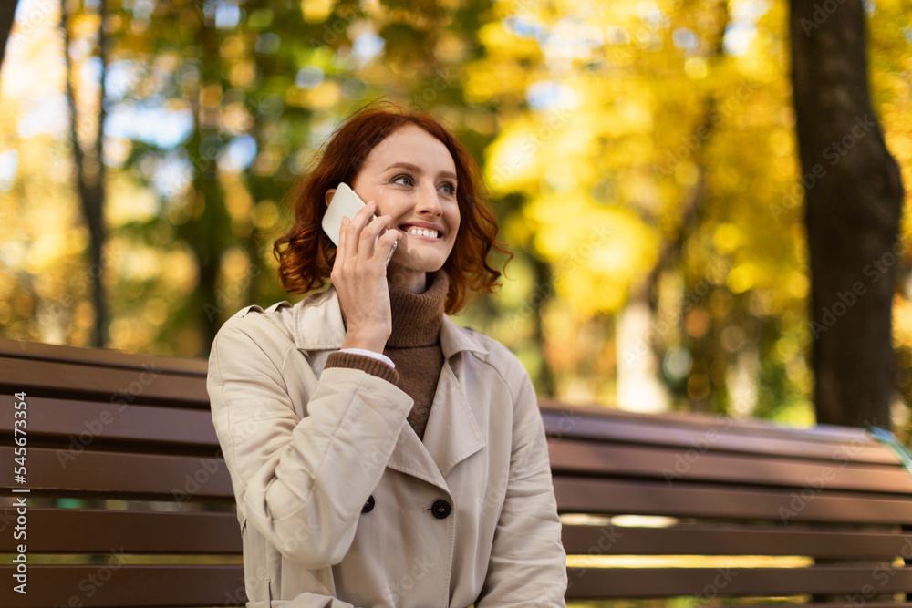 Cheerful pretty caucasian millennial woman with red hair in raincoat calling by phone, enjoy autumn and walk