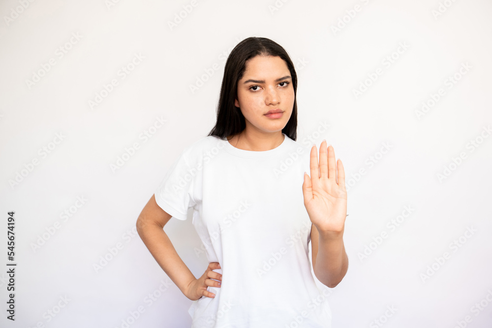 Portrait of serious young woman making stop gesture over white background. Caucasian lady wearing white T-shirt showing refusal sign and looking at camera. Forbiddance concept