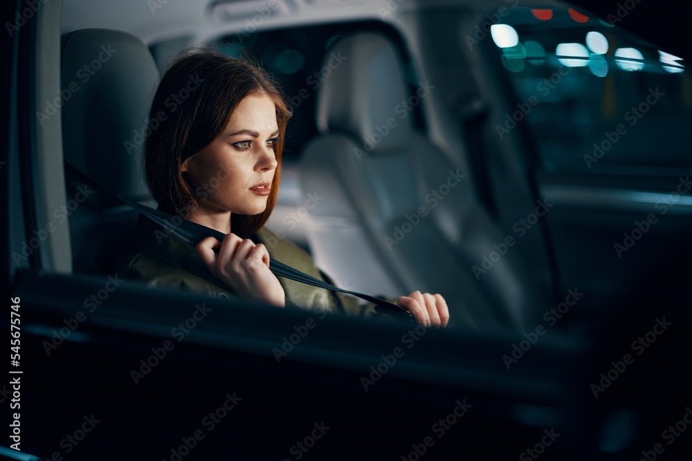 a sweet, relaxed, stylish woman is sitting in a black car in the passenger seat, at night and wearing a seat belt. Topics of road safety - obrazy, fototapety, plakaty 