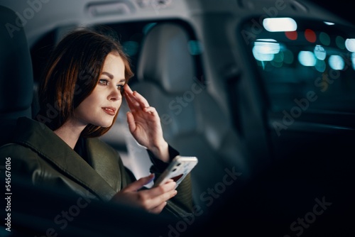  stylish, luxurious woman in a leather coat sitting in a black car at night on the passenger seat, looking thoughtfully to the side, holding her hand near her face, and the other holding a smartphone © SHOTPRIME STUDIO