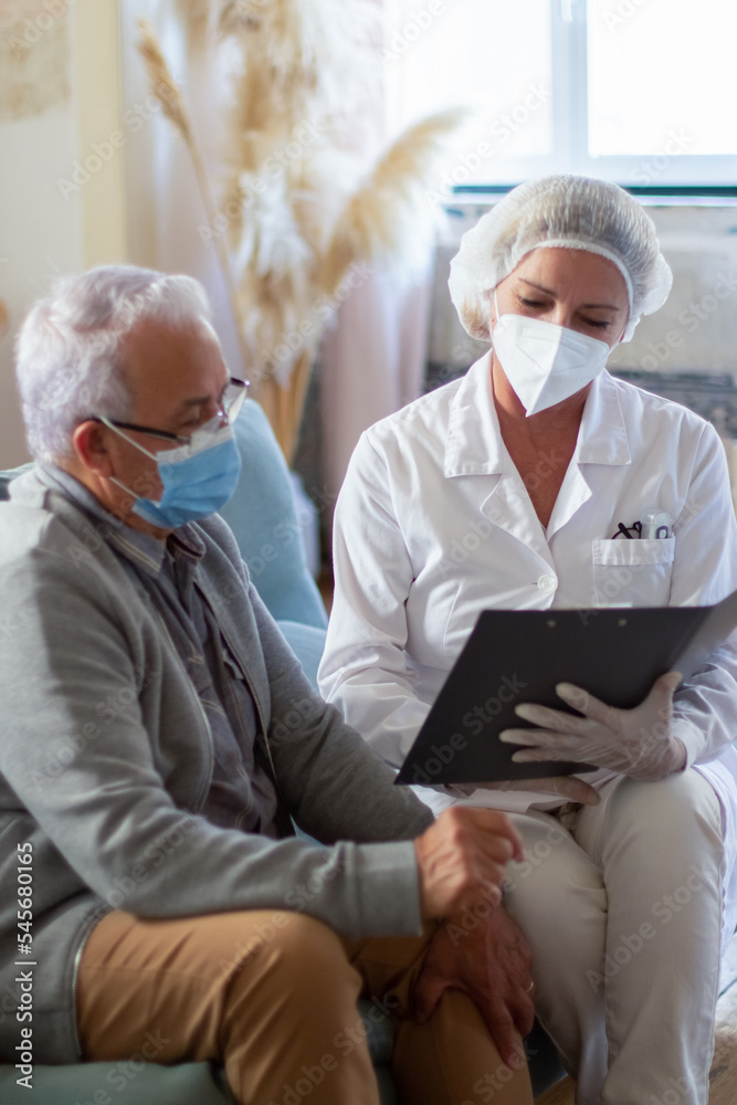 Obraz premium Portrait of female doctor at senior mans home during pandemic. Woman in medical uniform and man sitting on sofa, talking of vaccination before injection. Vaccination during virus diseases concept