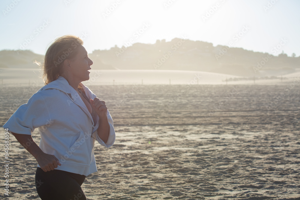 Side view of senior woman running on sandy beach in summer. Caucasian woman concentrated on training jogging and strengthening her health in morning. Sport, healthy lifestyle of elderly people concept