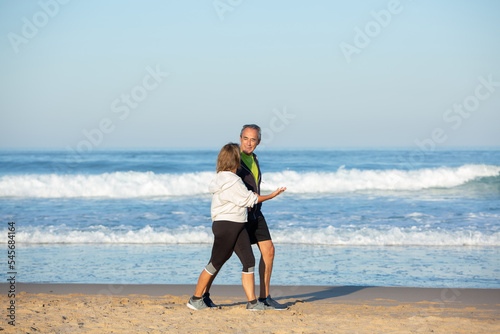 Cheerful senior couple talking while walking along shore. Long shot of beautiful mature man and woman in sportswear spending time together, looking at each other with tenderness. Communication concept