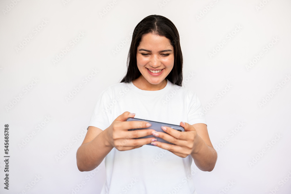 Portrait of happy young woman playing game or watching video on mobile phone. Caucasian lady wearing white T-shirt using smartphone over white background. Mobile technology concept Portrait of happy young woman playing game or watching video on mobile phone. Caucasian lady wearing white T-shirt using smartphone over white background. Mobile technology concept