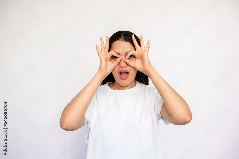 Portrait of surprised young woman making glasses with fingers. Caucasian lady wearing white T-shirt peeking through hands over white background. Astonishment and awe concept Portrait of surprised young woman making glasses with fingers. Caucasian lady wearing white T-shirt peeking through hands over white background. Astonishment and awe concept