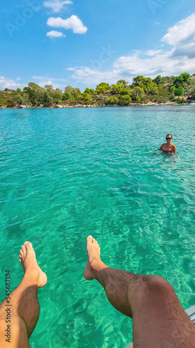 Feet of man hanging from bow of boat watching woman swimming in the blue lagoon of Vourvourou on peninsula Sithonia, Chalkidiki (Halkidiki), Greece, Europe. Tropical crystals clear turquoise water photo