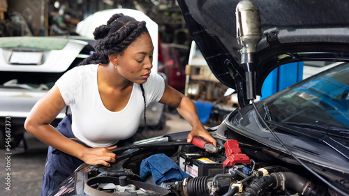 Charge electric power to ca battery by charging jumper cables. African mechanic female uses multimeter voltmeter to check voltage level in car battery at car service and maintenance garage.