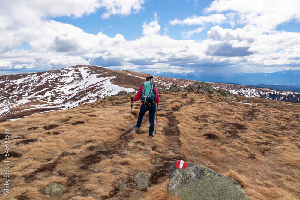 Woman walking next to path mark with Austrian flag painted on rock on remote alpine meadow near Ladinger Spitz, Saualpe, Carinthia, Austria, Europe. Hiking trail in the Austrian Alps in early spring