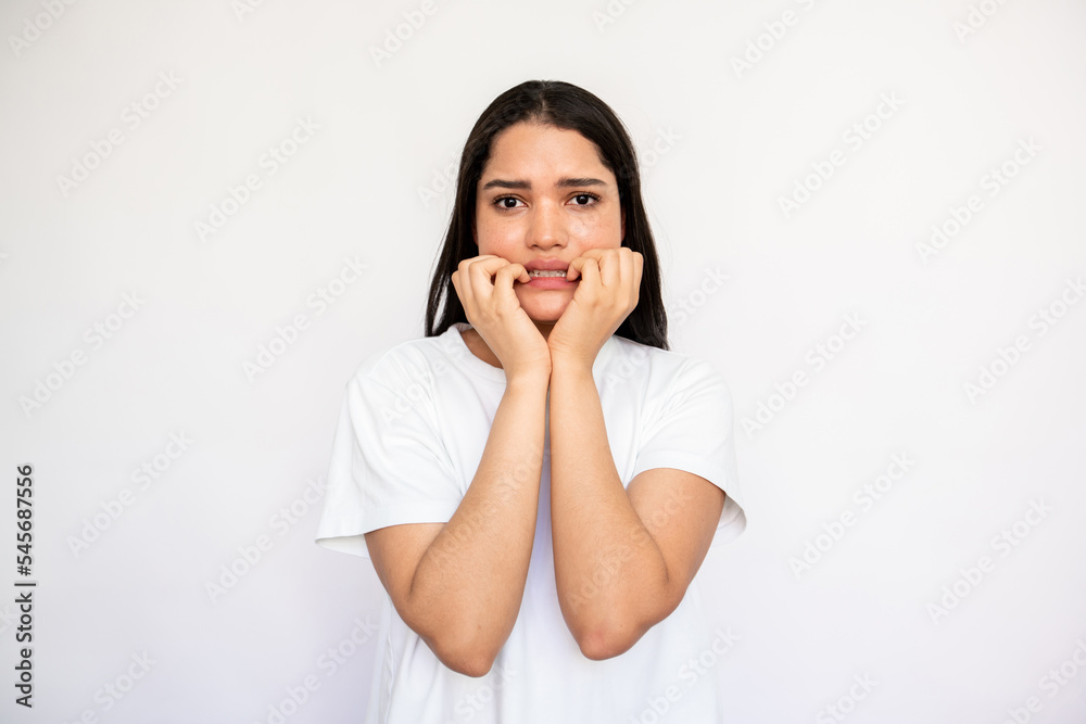 Portrait of neurotic young woman biting nails over white background. Caucasian lady wearing white T-shirt looking at camera in anxiety or fear. Stress and depression concept Portrait of neurotic young woman biting nails over white background. Caucasian lady wearing white T-shirt looking at camera in anxiety or fear. Stress and depression concept