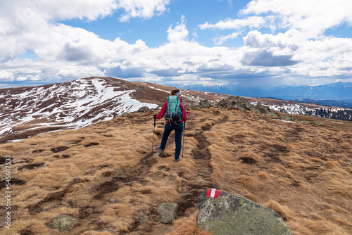 Woman walking next to path mark with Austrian flag painted on rock on remote alpine meadow near Ladinger Spitz, Saualpe, Carinthia, Austria, Europe. Hiking trail in the Austrian Alps in early spring