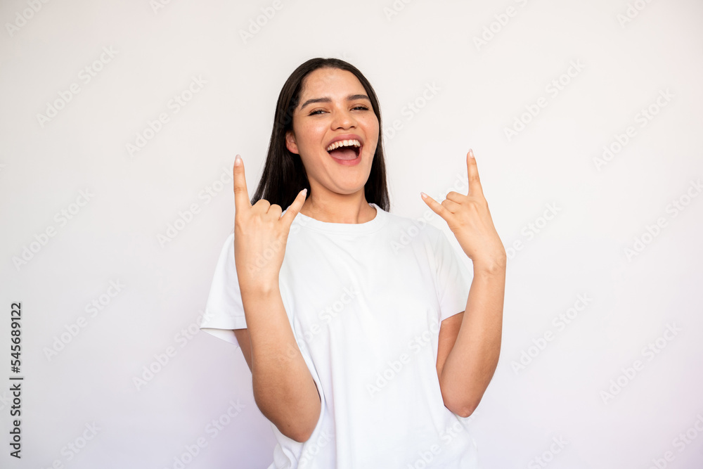 Portrait of excited young woman making rock and roll gesture over white background. Caucasian lady wearing white T-shirt looking at camera and laughing. Music fan concept Portrait of excited young woman making rock and roll gesture over white background. Caucasian lady wearing white T-shirt looking at camera and laughing. Music fan concept