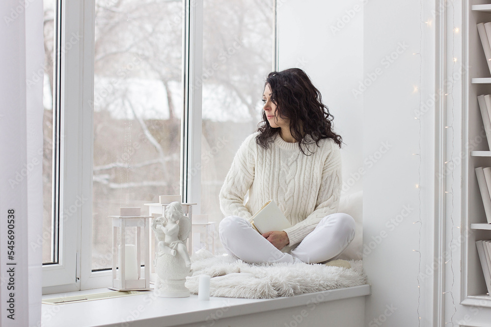BBrunette girl in white clothes sits near the window in a white room BBrunette girl in white clothes sits near the window in a white room