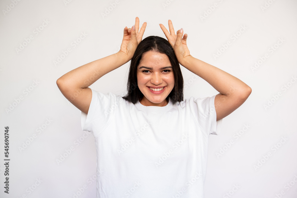Portrait of cheerful young woman making bunny ears over white background. Caucasian lady wearing white T-shirt having fun. Humor and fun concept Portrait of cheerful young woman making bunny ears over white background. Caucasian lady wearing white T-shirt having fun. Humor and fun concept