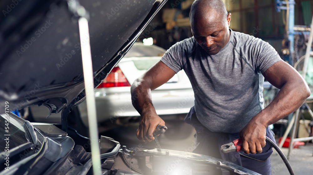 African american mechanic man uses  multimeter voltmeter to check voltage level in car battery at car service and maintenance garage.