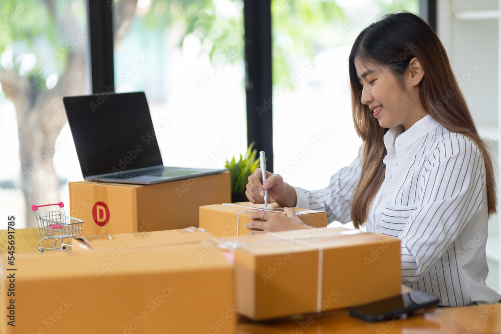 Woman in a small start-up business woman working in a home office working on a laptop and smartphone checking orders from the internet in preparation for shipping. Woman in a small start-up business woman working in a home office working on a laptop and smartphone checking orders from the internet in preparation for shipping.