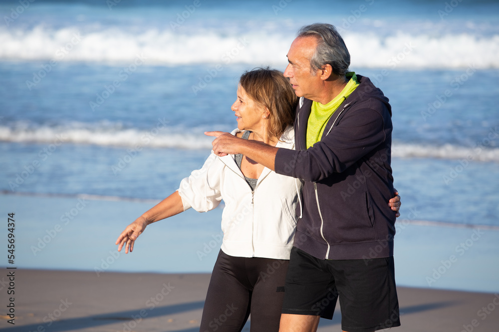 Senior couple hugging on seashore and looking to side. Medium shot of beautiful Caucasian mature man and woman in sportswear spending summer holiday at sea, exploring area. Love, sport concept Senior couple hugging on seashore and looking to side. Medium shot of beautiful Caucasian mature man and woman in sportswear spending summer holiday at sea, exploring area. Love, sport concept