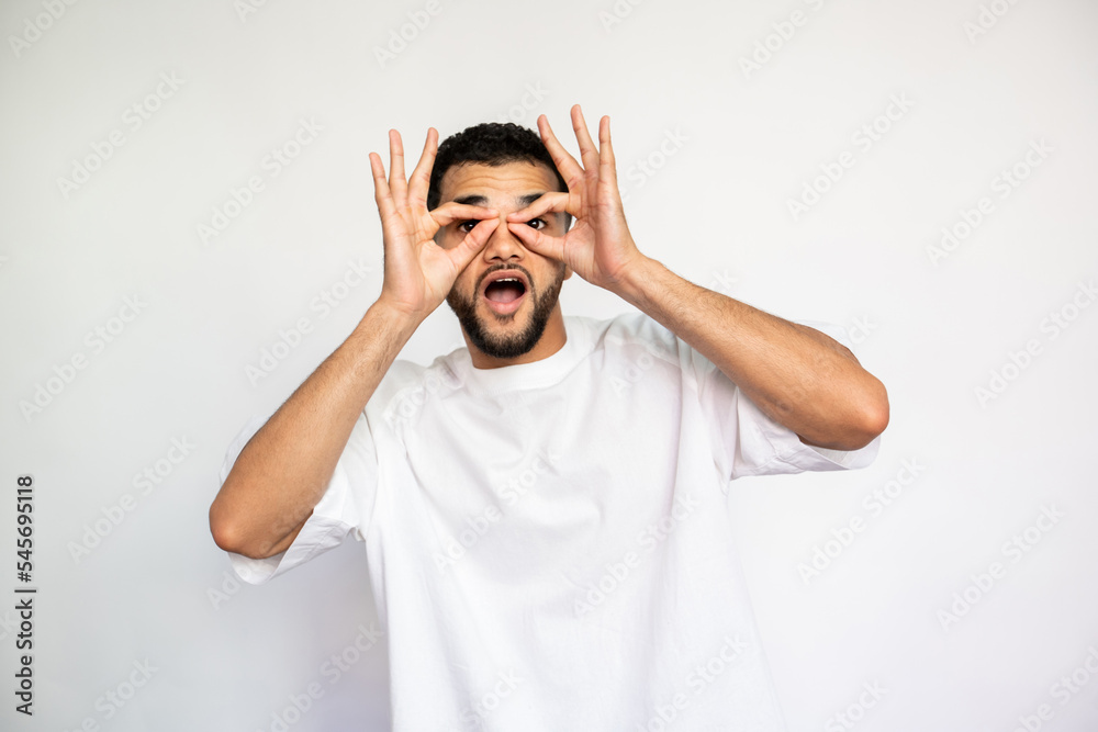 Portrait of excited young man doing glasses gesture. Amazed Caucasian male model with dark hair in white T-shirt looking at camera with open mouth and ok signs. Entertainment, grimacing concept Portrait of excited young man doing glasses gesture. Amazed Caucasian male model with dark hair in white T-shirt looking at camera with open mouth and ok signs. Entertainment, grimacing concept