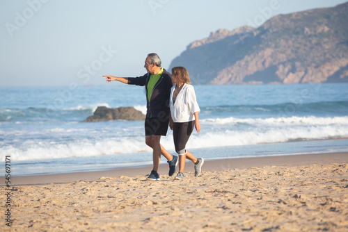 Man pointing finger into distance, walking on shore with wife. Long shot of cheerful senior couple spending time together, relaxing after cardio training, admiring seascape. Fitness, rest concept