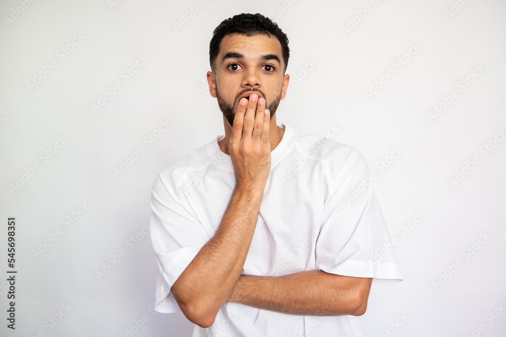 Portrait of amazed young man covering mouth with hand. Bored Latin American guy wearing white T-shirt looking at camera and yawning over white background. Astonishment and boredom concept Portrait of amazed young man covering mouth with hand. Bored Latin American guy wearing white T-shirt looking at camera and yawning over white background. Astonishment and boredom concept