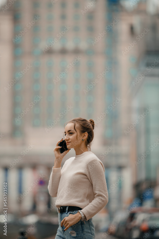 Fototapeta premium Young woman using mobile phone walking on the city street in Warsaw, Poland. Successful woman calling with cell telephone outside,business talking via mobile phone and walking on the city street.