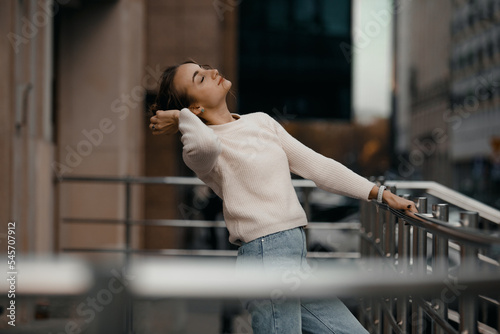 Professional woman worker wear standing outdoor terrace with a smile leaning on railing enjoy view from modern office skyscraper. Tall stylish girl near the railing and steps on the city background.