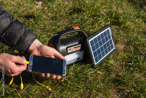 The man holds the phone in his left hand and puts it on the charger from a portable power station powered by a solar panel. A power station with a flashlight and a board powered by a solar panel. photo