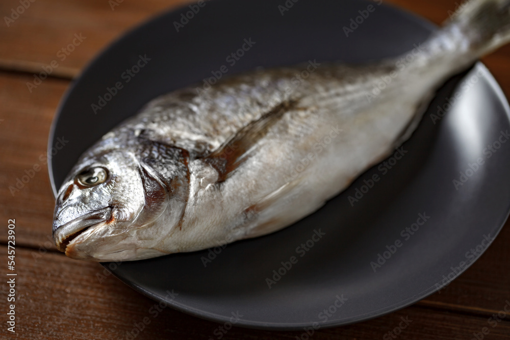 Dorado, raw fresh uncooked fish, on gray plate, on wooden brown plank tabletop background, side view, selective focus. Dorado, raw fresh uncooked fish, on gray plate, on wooden brown plank tabletop background, side view, selective focus.