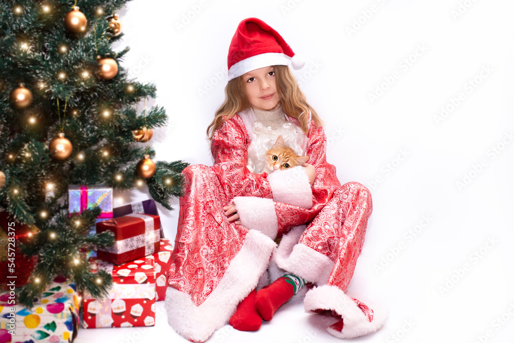 cute girl near christmas tree. a beautiful girl dressed as Santa Claus hugs a ginger cat near the Christmas tree. a child in a Christmas hat plays at home near the Christmas tree.