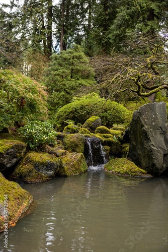 Vertical shot of a beautiful Japanese-style lush garden with moss-covered stones and a pond