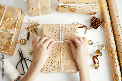 Merry Christmas! Hands wrapping stylish golden christmas gifts flat lay. Person preparing modern christmas gift boxes, festive paper, golden ornaments and thread composition on white wood photo