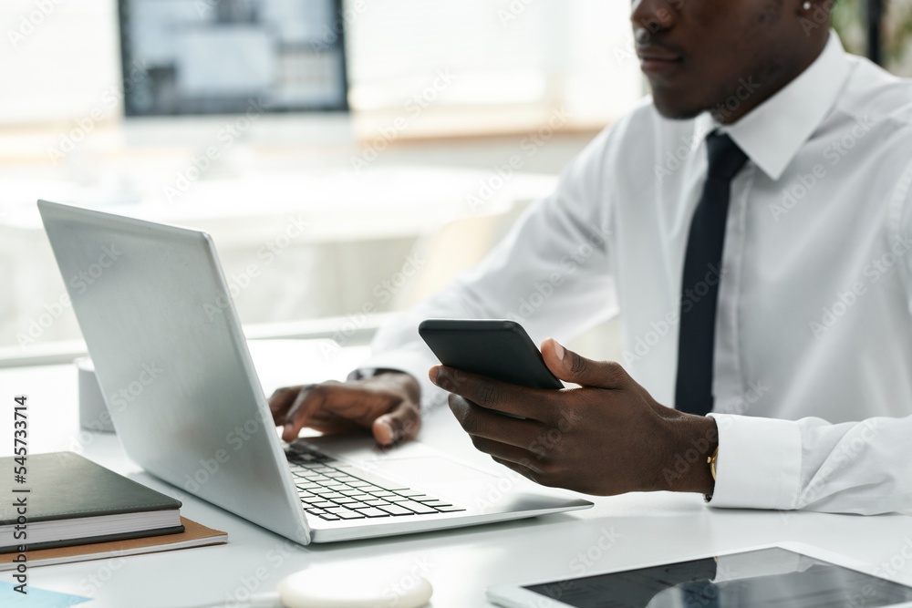 Young African American using smartphone and laptop at his work while sitting at table at office Young African American using smartphone and laptop at his work while sitting at table at office