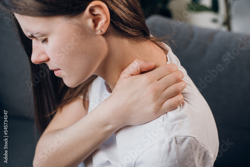 Close up of suffering sad young caucasian female touches and makes self-massage of upper back sitting on sofa at home, trapezius muscle and neck concept. Back pain, shoulder girdle spasm