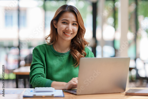 Attractive young Asian businesswoman using laptop in office smiling and looking at camera.