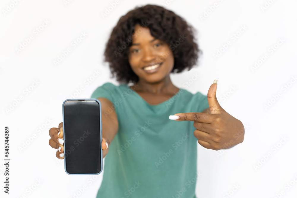 Pretty young woman pointing at smartphone. Smiling African American female model in blue T-shirt pointing at phone, advertising new application, close-up. Studio shoot, advertising concept Pretty young woman pointing at smartphone. Smiling African American female model in blue T-shirt pointing at phone, advertising new application, close-up. Studio shoot, advertising concept