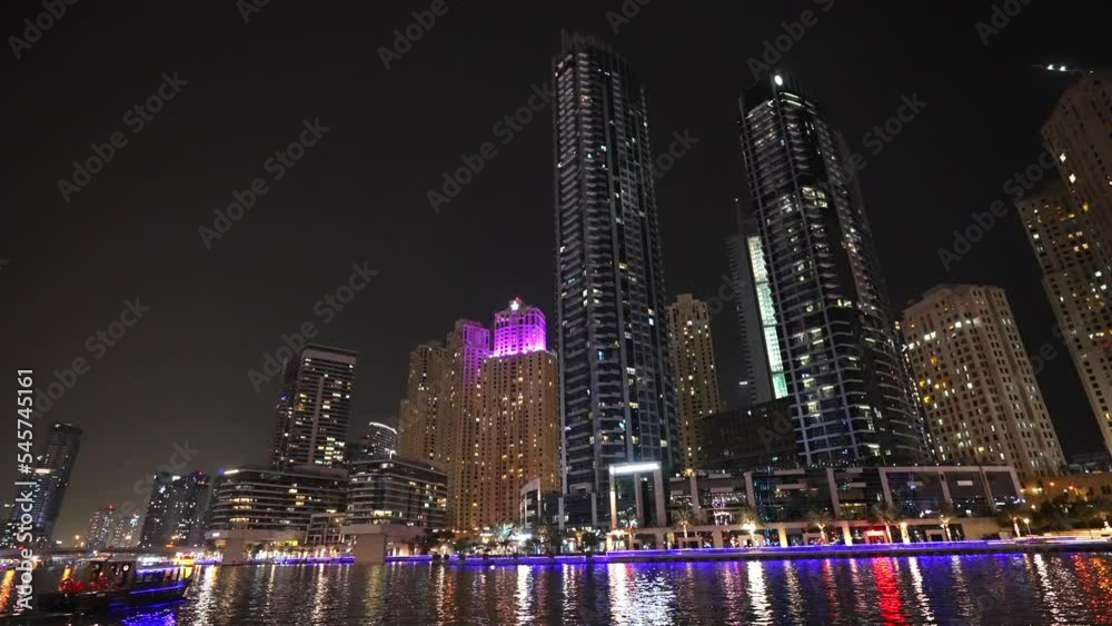 View of beautiful and developed city with skyscrapers at night. Buildings illuminated with neon color lights on seashore. Modern metropolis streets. Urban architecture. Reflection of lights in water.