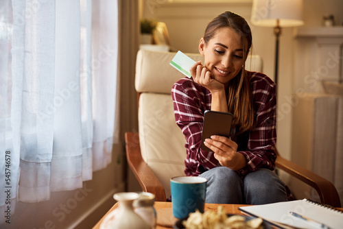 Happy woman online banking while using smart phone and credit card at home.