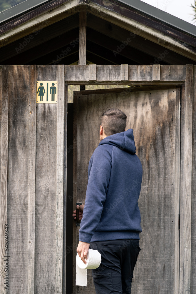 Young adult backpacker entering to rustic wooden toilet in outdoors background.