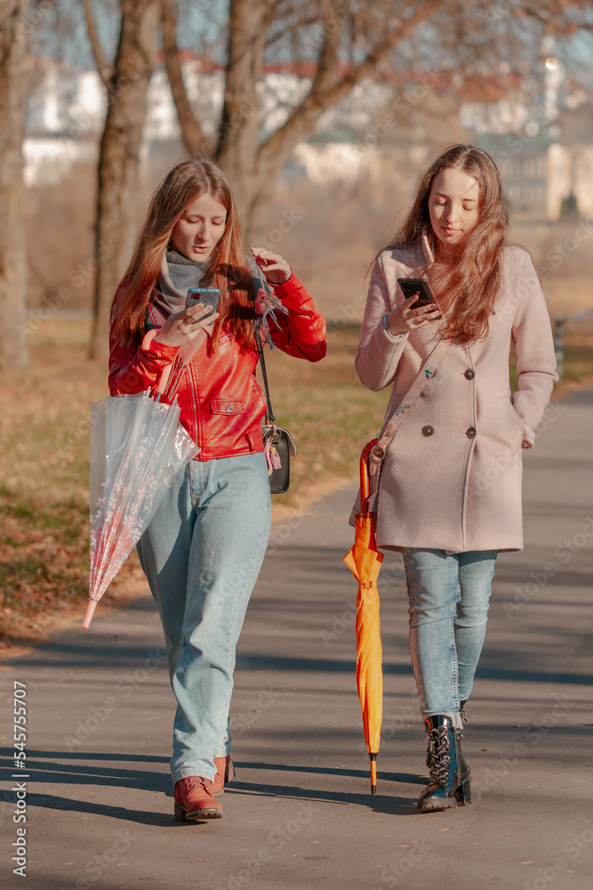 Two girls with umbrellas walk through the park in sunny autumn weather, holding smartphones in their hands and looking into them