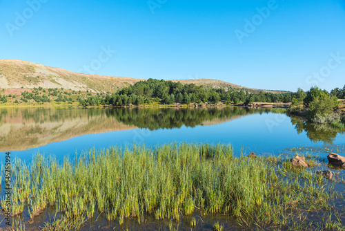Landscape of a lake surrounded by covered greenery mountains reflected in the water under a blue sky, Neila Lagoons Natural Park, Burgos, Spain