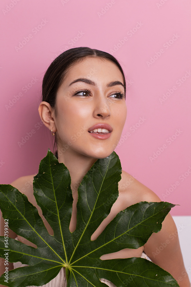Close up face of beautiful young eastern woman covering her face by green leaf on a pink background in the studio. Portrait of beauty woman with makeup and freckles standing behind big green leaves. Close up face of beautiful young eastern woman covering her face by green leaf on a pink background in the studio. Portrait of beauty woman with makeup and freckles standing behind big green leaves.