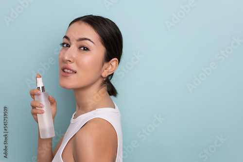 portrait of a beautiful young woman holding a bottle in her hand and spraying liquid with hydration on her face for rejuvenation on a blue background in the studio. © saulich84