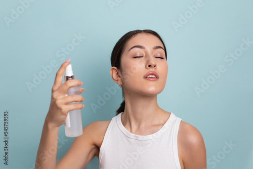 portrait of a beautiful young woman holding a bottle in her hand and spraying liquid with hydration on her face for rejuvenation on a blue background in the studio.