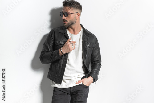 cool fashion bearded man with glasses looking to side, fixing jacket and posing