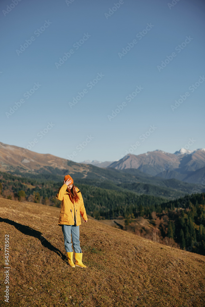 Full-length woman walking on a hill smiling with teeth and looking at the mountains in a yellow raincoat and jeans happy nature trip on a hike spring, freedom lifestyle 