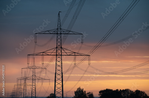 Close-up of large overhead power lines carrying electricity over long distances. The sun rises and shines in the background.