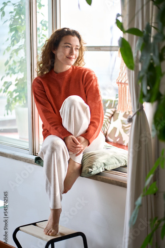 Happy serene young woman sitting on windowsill relaxing at home looking through window. Smiling calm lady chilling in apartment, dreaming, thinking of peaceful time enjoying peace of mind. Vertical