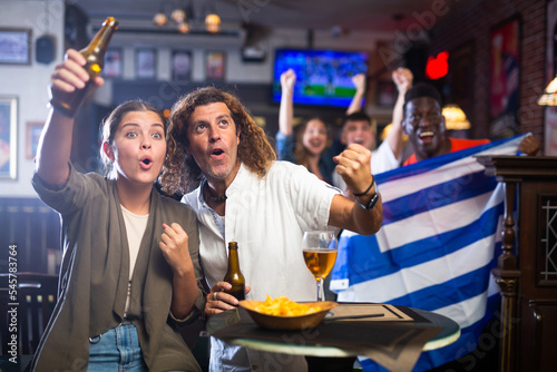 Happy multiracial soccer fans with flag of Greece celebrating victory of team with pint of beer and chips in the pub