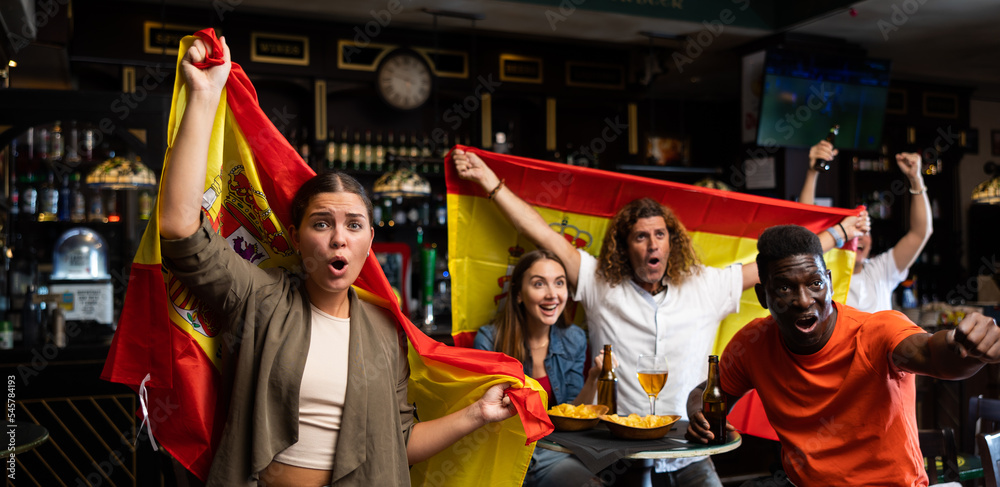 Emotional young girl waving national flag of Spain while watching football match in sports bar, cheering for favorite team and celebrating victory